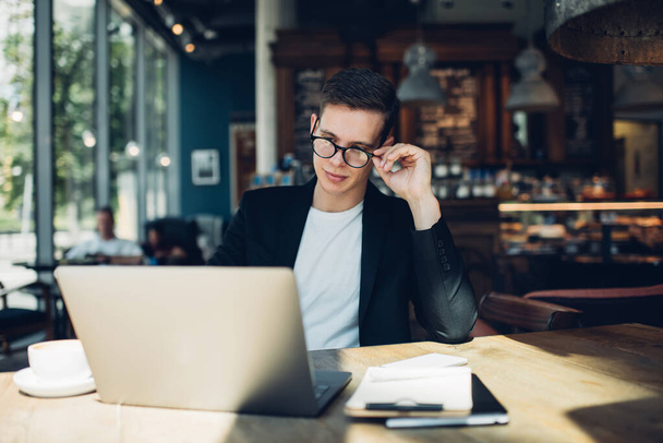 Улыбающийся молодой бизнесмен в очках и формальной одежде сидит за деревянным столом, настраивая очки и просматривая ноутбук во время работы над бизнес-проектом - Фото, изображение