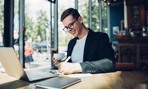 眼鏡をかけた幸せな若い男性起業家とカフェに座ってコーヒーを飲みながらメモを書きながらプロジェクトに取り組んでいる正式なスーツ - 写真・画像
