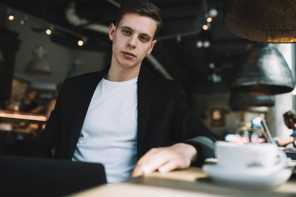 Confident young businessman sitting with hand on table and looking at camera while resting with cup of coffee in modern cafeteria - Foto, immagini