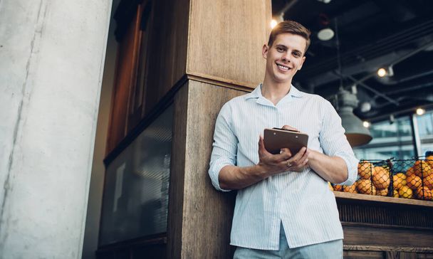 Smiling young man in casual clothes with clipboard leaning on counter in modern cafe outdoors and looking at camera while taking notes - Фото, зображення