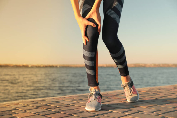 Young woman in sportswear having knee problems near river at sunset, closeup - Foto, immagini