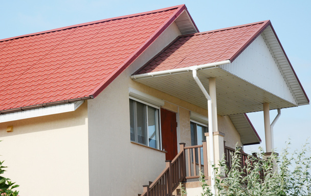 Un primer plano en una casa de estuco con un techo a dos aguas de azulejos de metal rojo con tablas de soffit y fascia, canaletas de lluvia con dos bajantes y un porche cubierto con escaleras de cubierta.  - Foto, Imagen