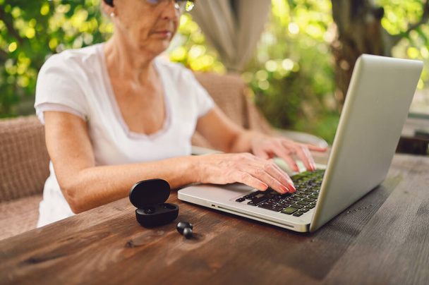テクノロジー、高齢者の概念-庭のラップトップコンピュータの屋外でオンラインで働く高齢者の高齢者の女性の手。前景のワイヤレスヘッドフォン。遠隔作業遠隔教育.  - 写真・画像