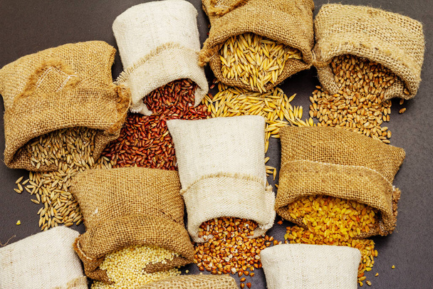 Variedad variada de cereales en sacos hechos a mano. Mezcla de ingredientes indispensables para una dieta saludable. Fondo de hormigón de piedra negra, primer plano - Foto, imagen