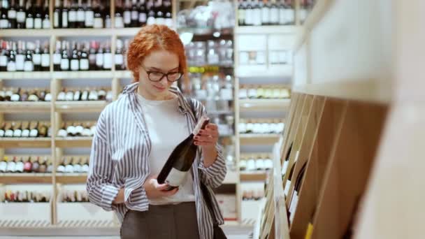 Vonzó vörös hajú nő figyelmesen választott egy üveg bort a modern élelmiszerboltban. Szupermarket koncepció - Felvétel, videó