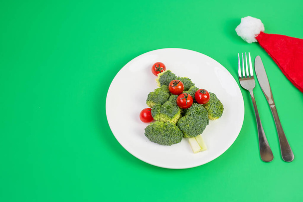 Рождественская елка из органических овощей на белой тарелке; Брокколи и помидоры с вилкой и ножом на зеленом фоне. Счастливого Рождества и с Новым годом, Новый Вы, здоровое питание и вегетарианская концепция - Фото, изображение