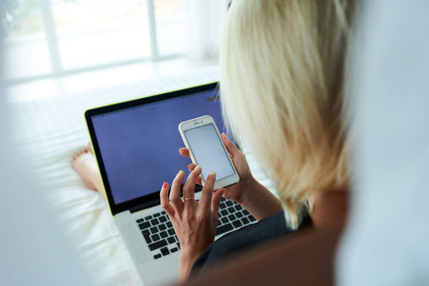 Back view of anonymous female with blonde hair checking messages on smartphone while lying on bed in bright room and chilling with laptop - Foto, Imagem