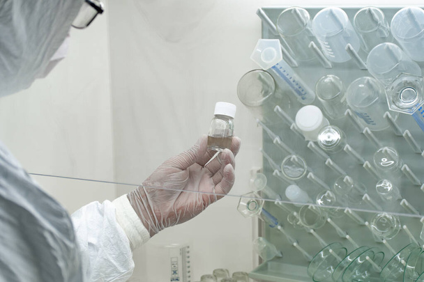 Scientist in protective white coat, mask and gloves analyzes a virus or bacteria sample in a laboratory with vials, glass flasks and chemicals. Medical lab test, new vaccine research or development - Fotó, kép