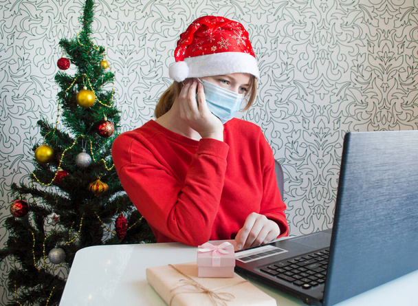 Sad young woman with medical mask and christmas presents, looking at laptop. Christmas atmosphere. Online communication - Zdjęcie, obraz