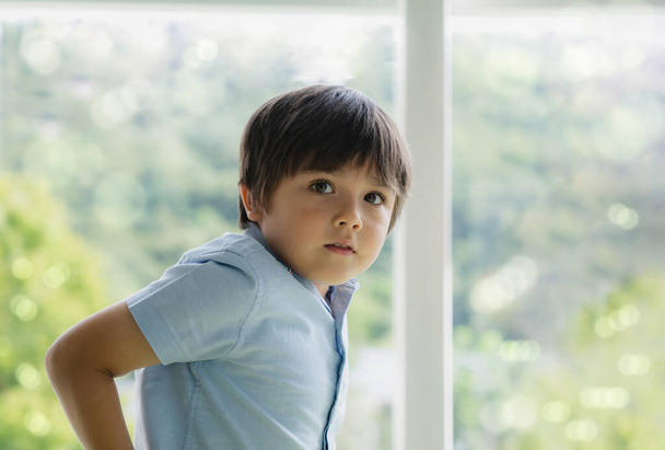 High-Key-Licht Portrait gutaussehendes Kind, das neben dem Fenster steht und mit lächelndem Gesicht in die Kamera schaut. Positive Kindheitserholung am Wochenende bei sonnigem Frühlings- oder Sommertag - Foto, Bild