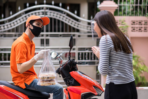 Mad food deliveryman με μάσκα προσώπου σε πορτοκαλί στολή περιμένετε για μοτοσικλέτα, ενώ η γυναίκα πελάτης συγγνώμη για να πάρει αρτοποιείο αργά. Πανδημία COVID-19. Νέο φυσιολογικό να χρησιμοποιήσετε την εφαρμογή smartphone για να παραγγείλετε. - Φωτογραφία, εικόνα