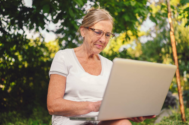 Tecnologia, conceito de pessoas de idade avançada - idosa feliz idosa trabalhando on-line com computador portátil ao ar livre no jardim. Trabalho à distância, educação à distância.  - Foto, Imagem