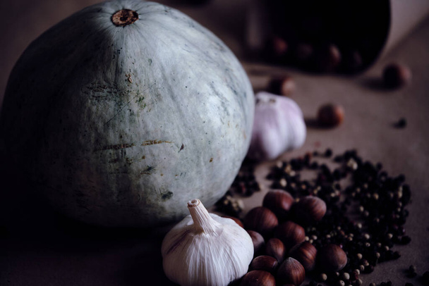 Blue pumpkin, Hazelnuts in nutshell, Fresh garlic bulbs with Mixed sorts and colors peppercorns on beige  background. Muted brightness image. Concept of autumnal harvest and organic vegetarian food. - Фото, зображення