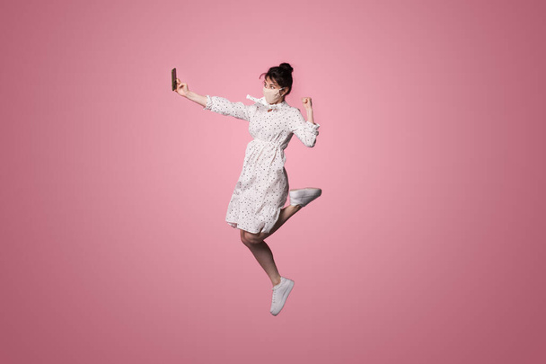Caucasian woman with a medical mask is wearing a dress and making selfie using a phone while jumping on a pink studio wall - Photo, Image