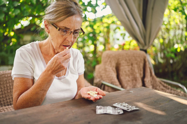 Starsza pani w okularach bierze tabletki witaminowe na zewnątrz w ogrodzie. Koncepcja opieki zdrowotnej dla osób starszych - Zdjęcie, obraz