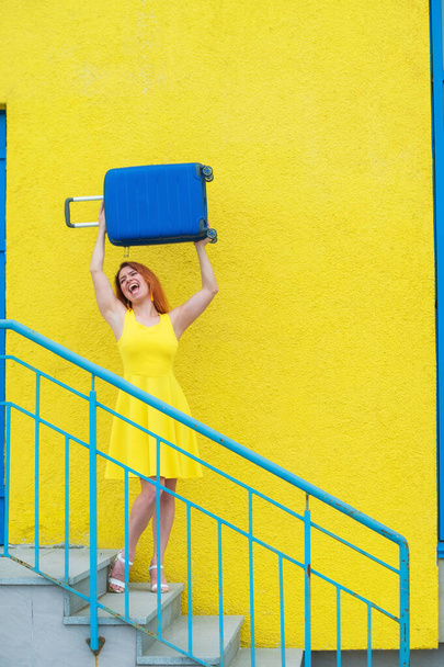 Beautiful red-haired smiling woman joyfully descends the stairs and waving a blue suitcase on a yellow background - Foto, Imagen