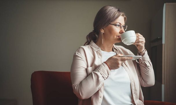 Foto vista lateral de una mujer mayor bebiendo una taza de té mientras mira a través de la ventana desde el interior de la casa - Foto, imagen