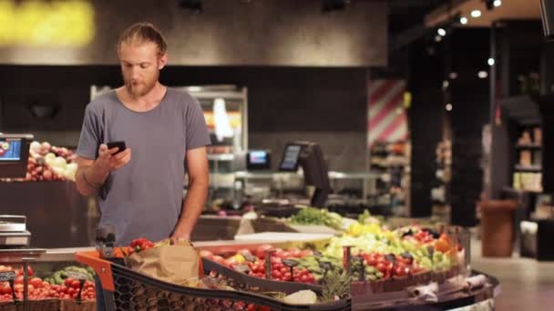 Attraktiver lässig blonder bärtiger Mann mit Einkaufswagen voller Waren unterhält sich beim Einkaufen im modernen Lebensmittelgeschäft intensiv auf dem Smartphone - Filmmaterial, Video