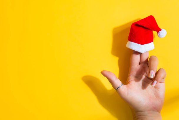Creative Christmas backdrop with woman 's hand and Santa Claus red hat on her fingers on a yellow background, copy space. Плоский лежал. Поздравительная открытка. - Фото, изображение