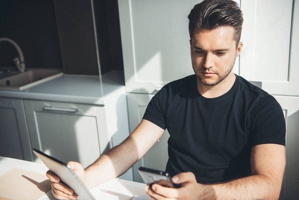Fehér üzletember dolgozik távolról otthon egy telefon és digitális tabletta pózol a konyhában - Fotó, kép