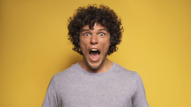 Emotional young man screams. Caucasian guy opens mouth bulging eyes while looking at camera. Isolated on yellow background - Foto, imagen