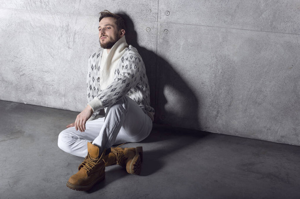 Perfil de um jovem confiante em jeans brancos e casaco de lã, sentado, posando sobre fundo de concreto. - Foto, Imagem