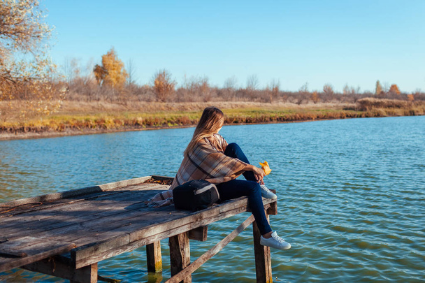 Путешественник с рюкзаком отдыхает на осенней реке на закате держа листья. Молодая женщина сидит на пирсе, любуясь пейзажем и погодой. Активный образ жизни - Фото, изображение