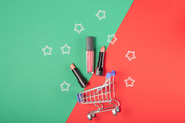 Women's lipsticks and lip glosses in a shopping trolley on a bright red and green background. The concept of buying cosmetics, online shopping, holiday - Foto, Imagem