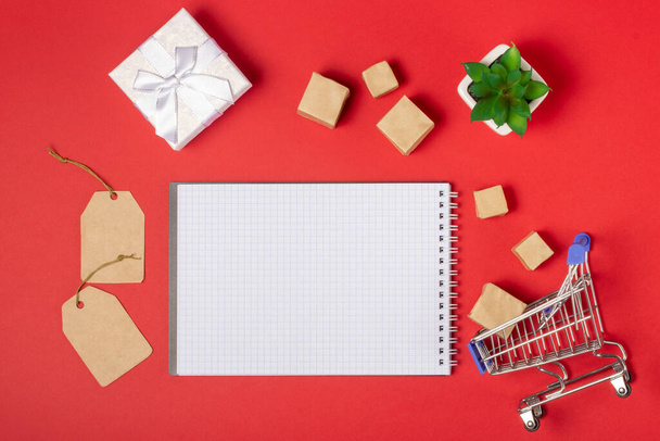 Open blank notebook with place to add text, shopping cart and boxes on a bright red background. Online shopping concept - Foto, Imagem