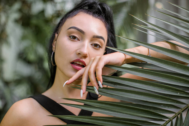 Summer fashion beauty portrait. Close up of face of beautiful Asian Japanese woman with long dark hair, wearing black dress, posing to camera with tropical palm trees and exotic plants on background - Foto, Imagem