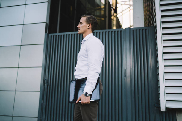 Side view sophisticated young man in white shirt and dark trousers standing in front of modern building with cased tablet in hand and looking away - Photo, image