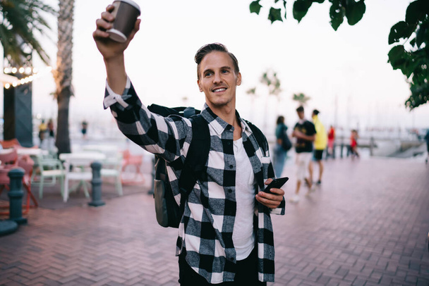 Happy caucasian hipster guy in trendy wear raising coffee cup offering beverage standing on street, smiling male traveler in casual outfit gesture and smiling exploring city on vacations on square - 写真・画像