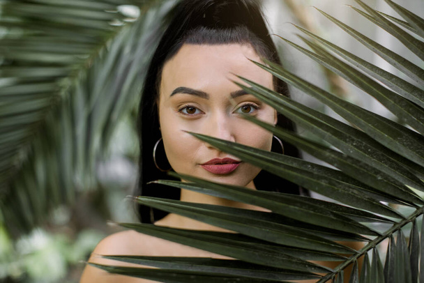 Natural beauty, exotic woman. Young pretty sexy Asian woman model with long black ponytail hair and natural make up, posing to camera behind tropical palm leaves. Close up portrait - Foto, Imagen