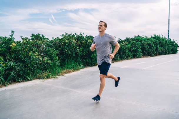 Muscular male athlete in electronic headphones have cardio training during morning workout in city, Caucasian pumped man in earphones running and listening motivation podcast during jogging - Fotoğraf, Görsel