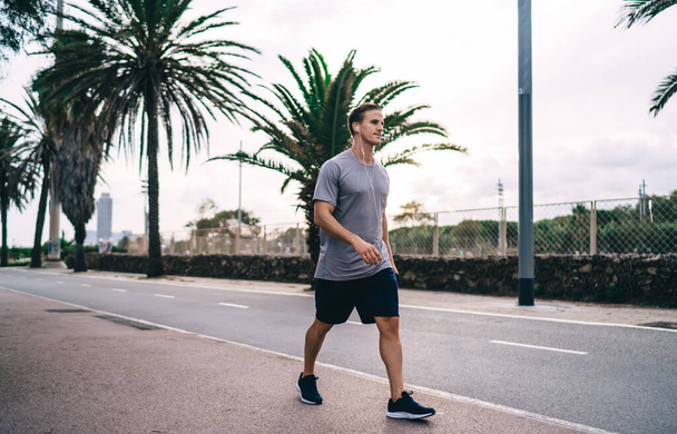 Caucasian male in electronic headphones walking at street using modern technology during morning time for training muscles, bodybuilder in earphones listening audio book with motivation plot - 写真・画像