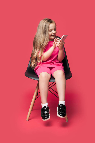 Blonde caucasian girl is sitting in a chair and smile using a phone ona red studio wall - Foto, Imagem