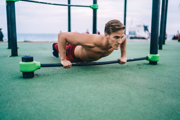 Young Caucasian male with muscular hands pushing up from bars at sports playground using equipment for warming up, sporty shirtless man enjoying actively exercising for keeping perfect body shape - Fotoğraf, Görsel