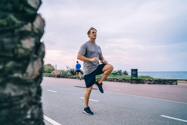 Energeting caucasian male runner warming muscles of legs jumping on street satisfied with vitality and physical strength,hipster guy spending free time actively having morning workout outdoors leaping - Fotoğraf, Görsel