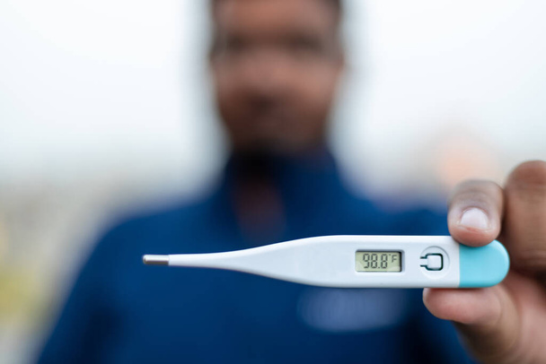 Hombre enfermo sostiene termómetro digital con lectura de temperatura corporal en Fahrenheit cerca de la cámara. - Foto, imagen