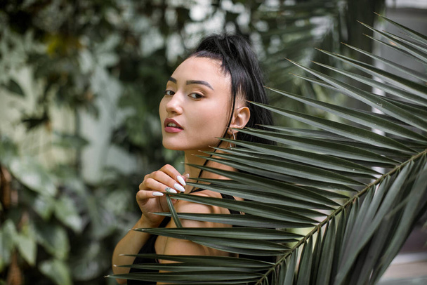 Portrait of beautiful sexy asian model with perfect face, wearing elegant black dress, posing to camera with palm tree leaf and exotic greenhouse plants on the background. Natural beauty - Zdjęcie, obraz