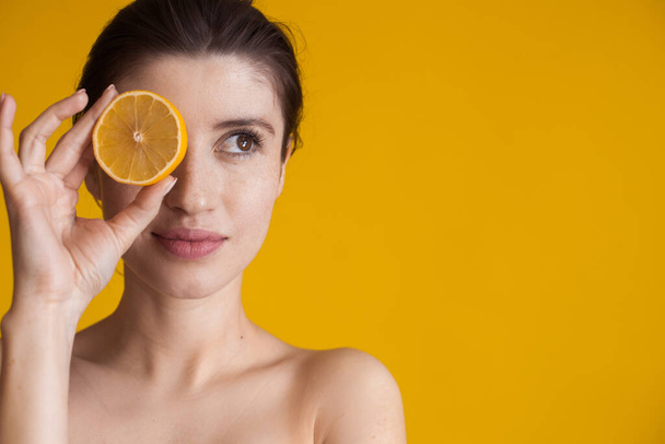 Mujer caucásica con la piel desnuda cubre el ojo con un limón durante el día de spa posando en una pared amarilla en el estudio - Foto, imagen