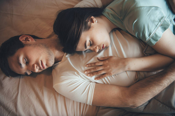 Brunette caucasian woman is sleeping on her boyfriend chest relaxing together in bed covered with a quilt - 写真・画像