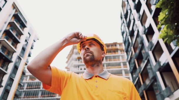 Operaio barbuto in uniforme arancione che indossa un casco sulla testa mentre si trova fuori dall'edificio durante la costruzione. caucasico maschio lavoratore edile in abiti da lavoro indossa un casco protettivo. - Foto, immagini