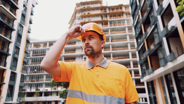 Construction worker putting on his safety gear helmet at job site. Safety first, profession concept. Builder puts on hard hat uniform and prepares for work. Man puts on cap orange construction site. - Photo, Image