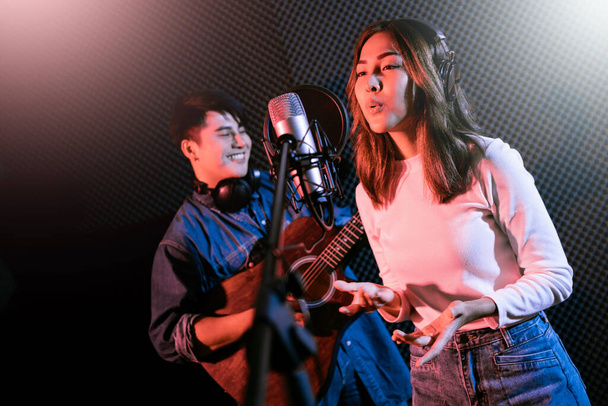 Pretty Asian female singer recording songs by using a studio microphone and pop shield on mic with male playing guitar in blue and red light. Performance and show in the music business. Duet session. - Valokuva, kuva