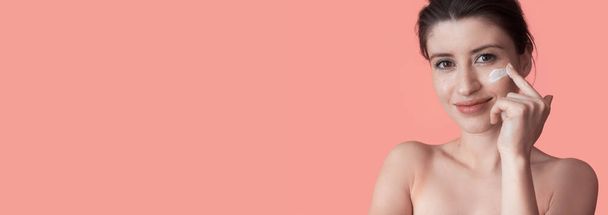 Η φακιδομένη γυναίκα με τους γυμνούς ώμους ποζάρει σε έναν τοίχο στούντιο με ελεύθερο χώρο απλώνοντας μια κρέμα στο πρόσωπό της και χαμογελώντας στην κάμερα - Φωτογραφία, εικόνα