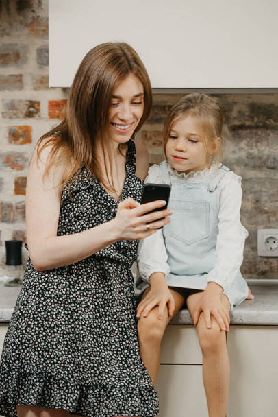 A European smiling young mother with pale skin is reading the news on a phone with a daughter in the kitchen. A mom is using a cellphone while her happy pretty girl is sitting on a countertop. - Fotoğraf, Görsel