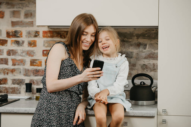 Eine junge europäische Mutter mit blasser Haut lacht, während sie mit einer Tochter in der Küche die Nachrichten am Telefon liest. Eine Mutter benutzt ein Handy, während ihr hübsches Mädchen auf einer Arbeitsplatte sitzt. - Foto, Bild