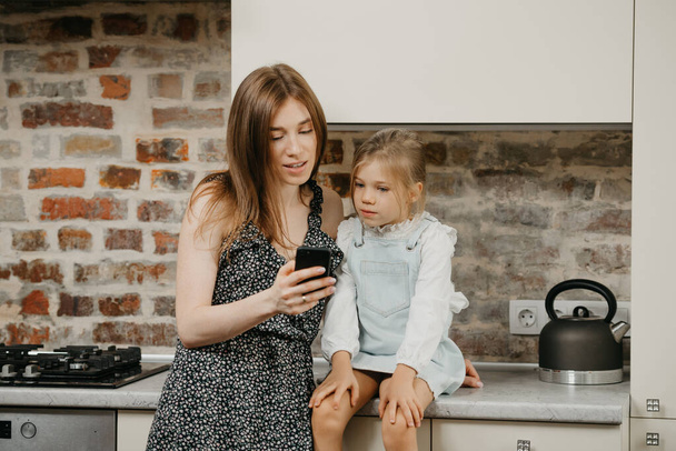 Eine junge europäische Mutter mit blasser Haut liest die Nachrichten am Telefon mit einer Tochter in der Küche. Eine Mutter benutzt ein Handy, während ihr hübsches Mädchen auf einer Arbeitsplatte sitzt. - Foto, Bild