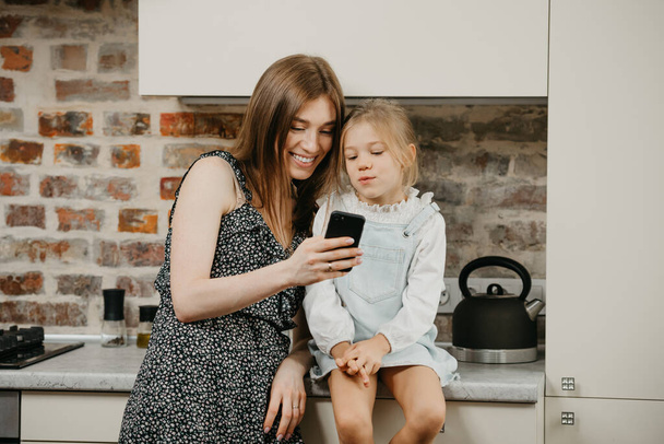 A European young mother with pale skin is smiling while reading the news on a phone with a daughter in the kitchen. A mom is using a cellphone while her pretty girl is sitting on a countertop. - Foto, afbeelding